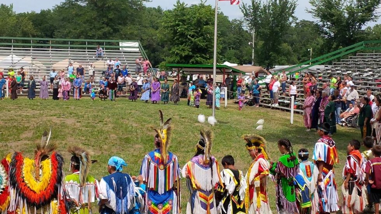 Group of individuals in traditional native clothing standing in a circle in an open green space.