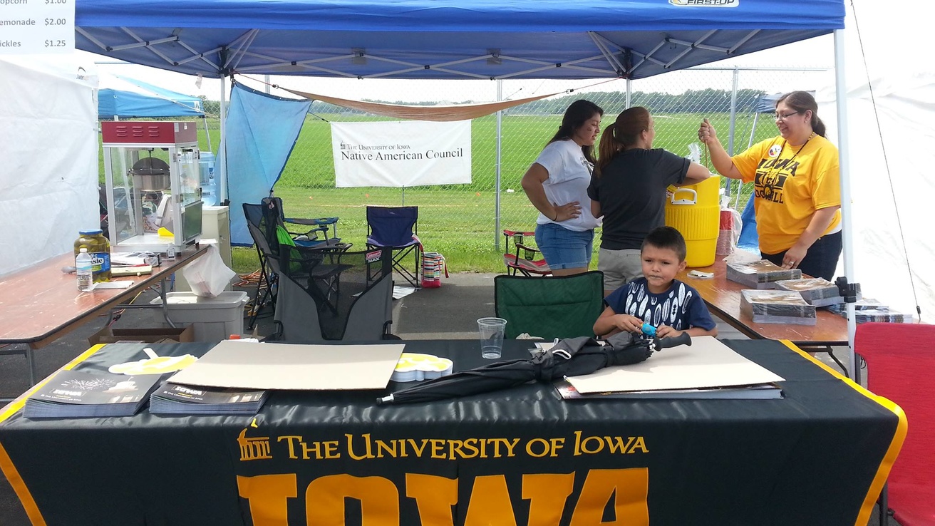 Blue tent with table in front with yellow and black "IOWA" tablecloth. Three individuals standing in back near water cooler.
